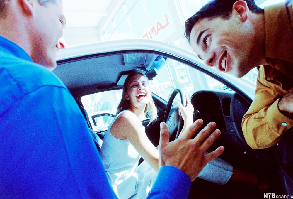 En kvinne sitter bak rattet i en bil og kikker ut av den åpne framdøra, der to menn står rett utenfor. De tre er i en samtalesituasjon og smiler og ler. Foto.