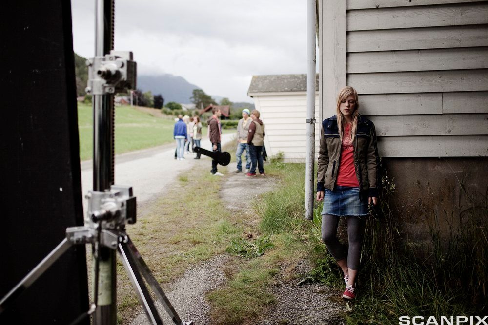 Ei ung jente står bak eit hushjørne og ser ut til å gøyme seg, mens ungdom samtaler i bakgrunnen. Foto.