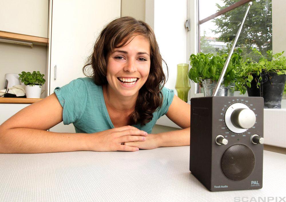 Foto av en smilende kvinne som lener seg ned på et kjøkkenbord der det står en radio.