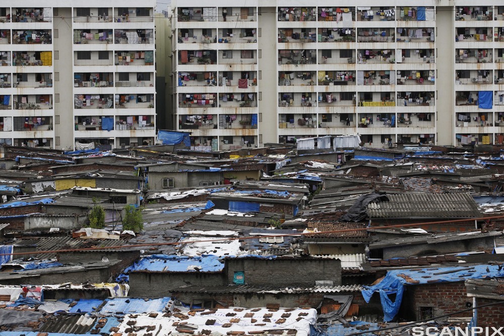 Lave hus med tak av bølgeblikk og plast i forgrunnen og boligblokker der de fleste leilighetene har klær som henger til tørk, på balkongene. Foto.