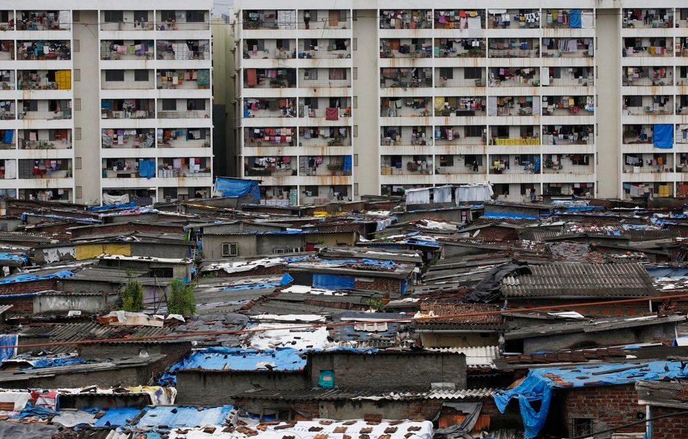 Lave hus med tak av bølgeblikk og plast i forgrunnen og boligblokker der de fleste leilighetene har klær som henger til tørk, på balkongene. Foto.