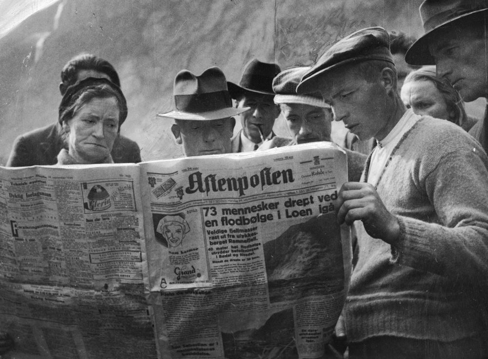 Flere mennesker leser ei avis de holder opp foran seg. Overskriften på forsiden er "73 mennesker drept ved en flodbølge i Loen i går". Svart-hvitt-foto.