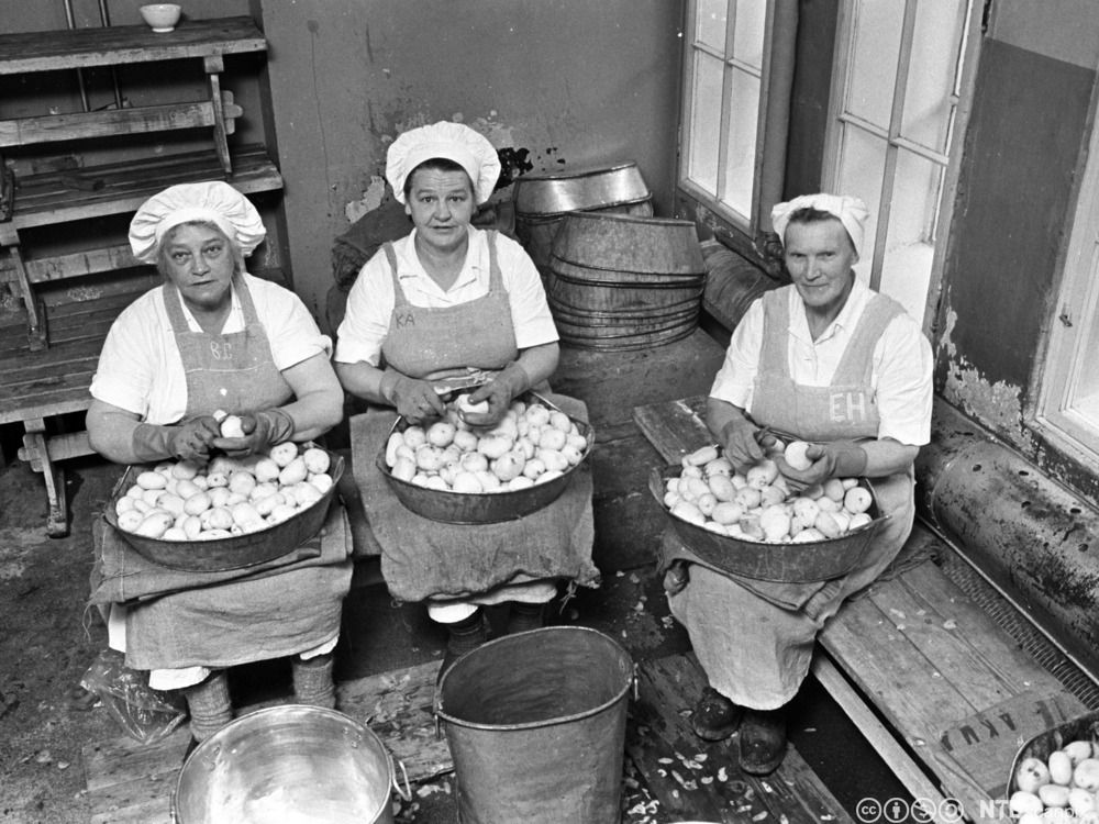Tre kvinner med forkle og hvite hodeplagg skreller poteter. De har hvert sitt kar i fanget hvor de legger de ferdigskrelte potetene. Svart-hvitt foto.