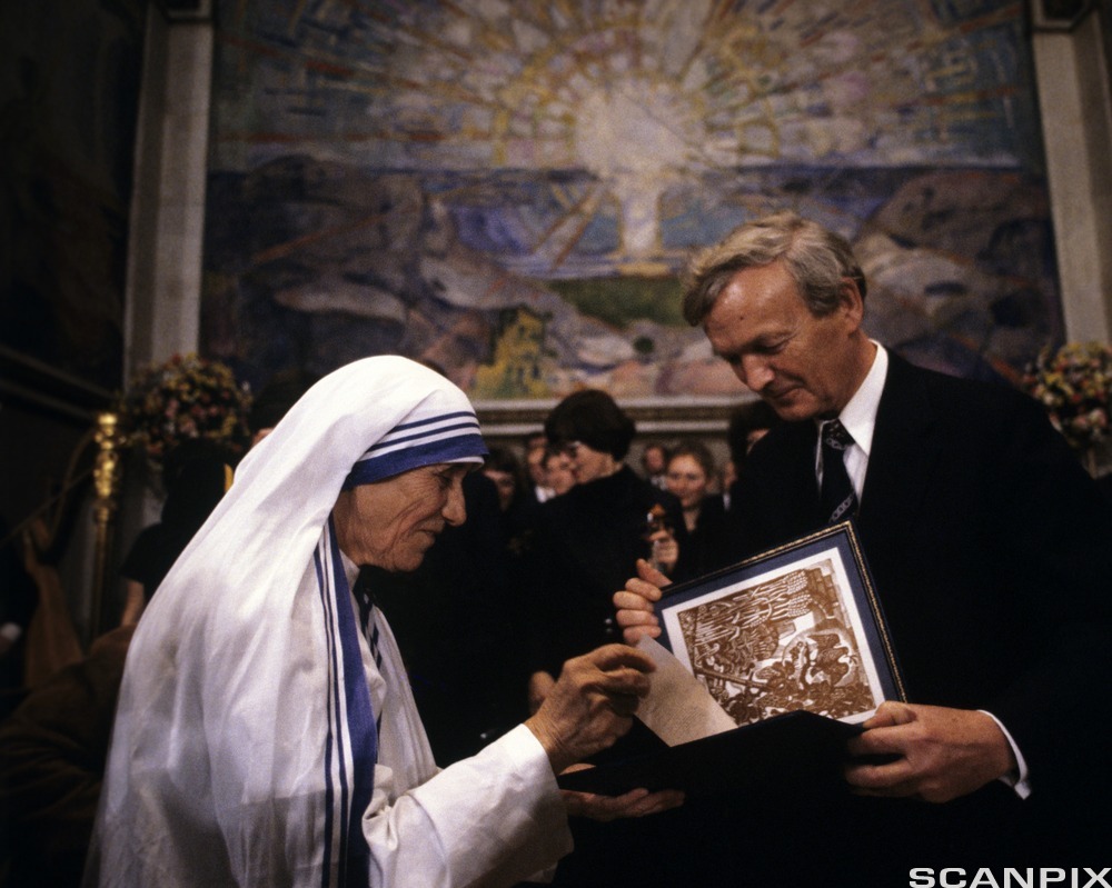 Eldre kvinne i hvit drakt og hvitt hodeplagg og en eldre mann i dress holder begge i og ser ned i et omslag med en illustrasjon inni. I bakgrunnen er det flere mennesker samlet og bakerst er et stort fargerikt veggmaleri. Foto.