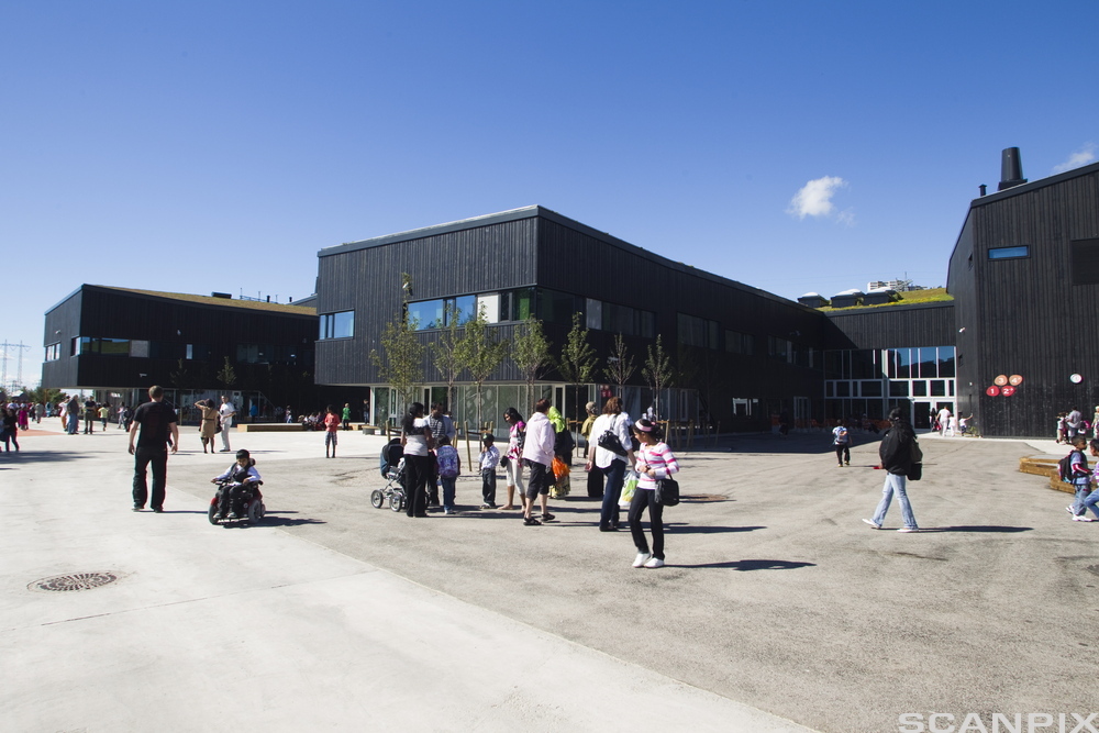 Åpen plass foran en stor bygning. Flere personer står og går på plassen. Foto.
