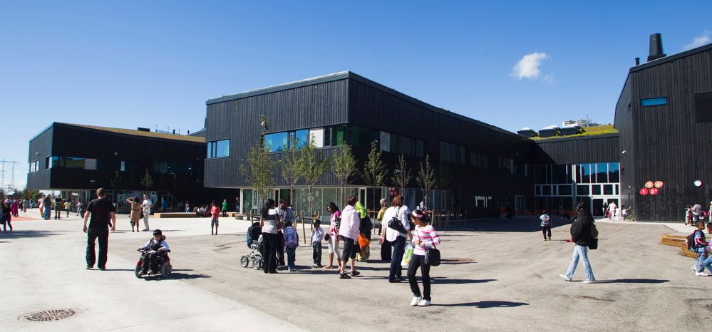 Åpen plass foran en stor bygning. Flere personer står og går på plassen. Foto.