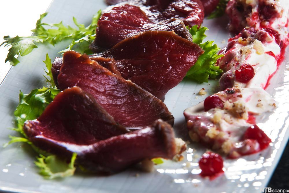 Tynne kjøttstykker anrettet med grønn salat, røde bær og rømme på hvit tallerken. Foto.