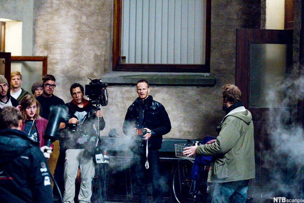 Stemningsbilde fra et filmsett. To menn står i en bakgård, det er røyk i lufta. Rundt står flere filmarbeidere med blant annet kameraer og mikrofoner. Foto.