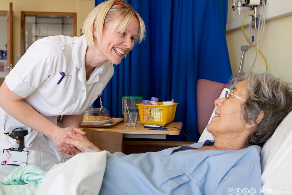 Kvinnelig helsefagarbeider smiler og holder en eldre kvinnelig pasient i hånda. Pasienten ligger i senga, hun smiler også. Foto.
