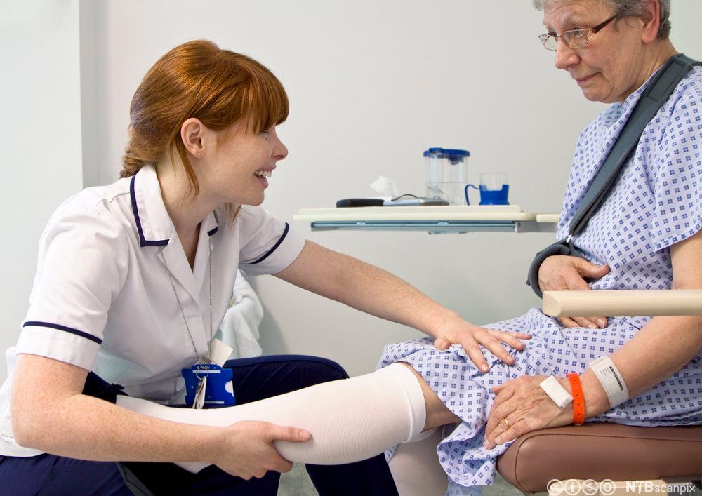 Kvinnelig pasient får hjelp av kvinnelig helsefagarbeider til å ta på støttestrømper. Foto.