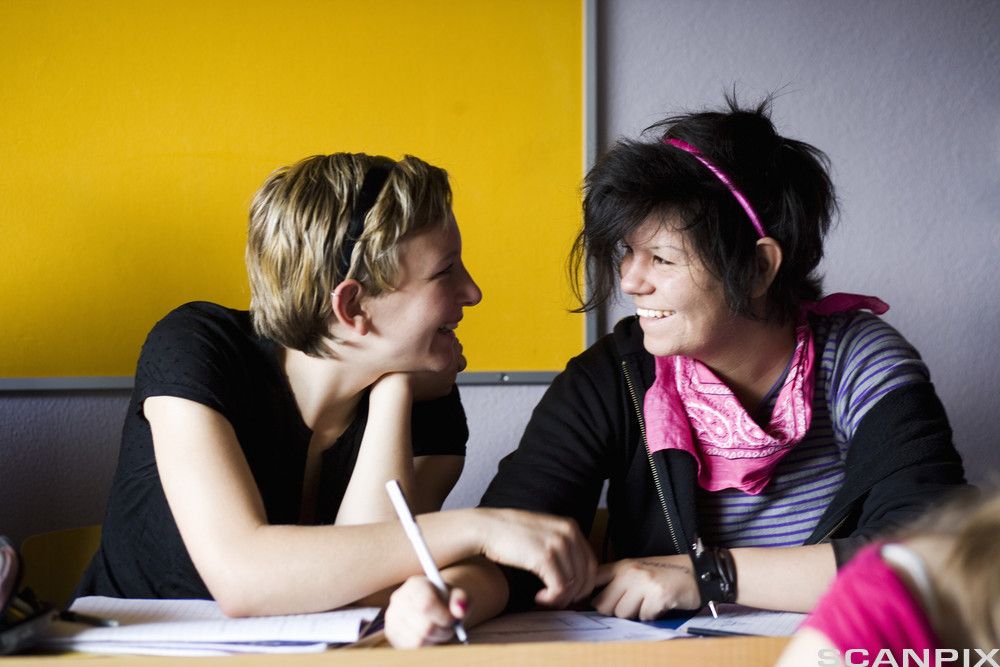 To jenter sitter ved et bord med notatbøker på. Jentene smiler mot hverandre, mens den ene skriver noe i notatboka. Foto.