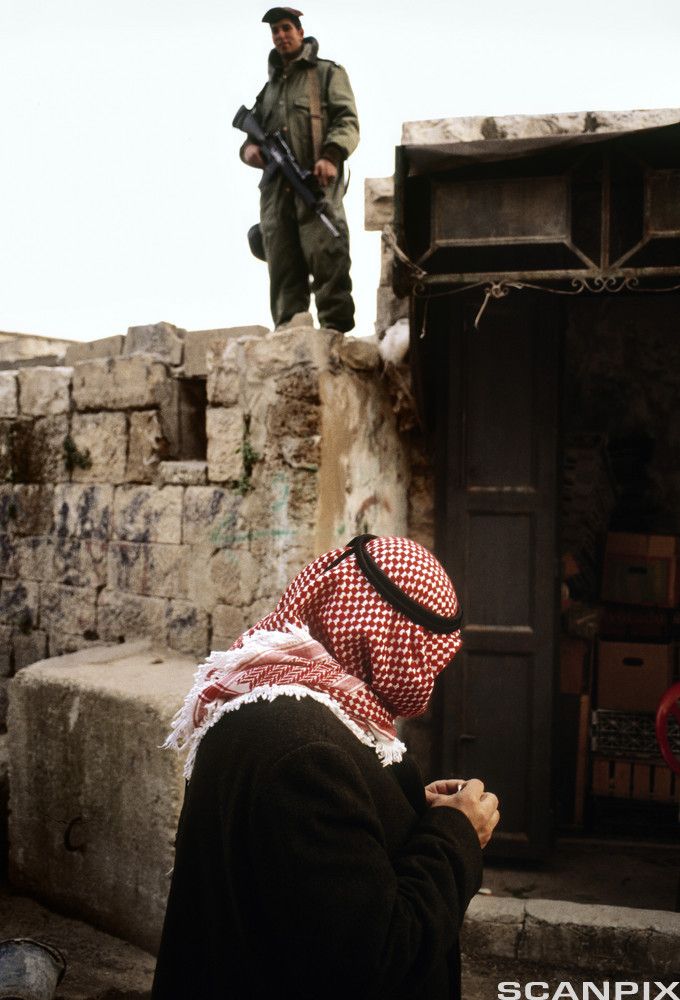 En ung soldat med maskingevær står på toppen av en mur mens en eldre mann med rødt palestinaskjerf på hodet passerer i gata under han. Bildet er tatt i froskeperspektiv. Foto.
