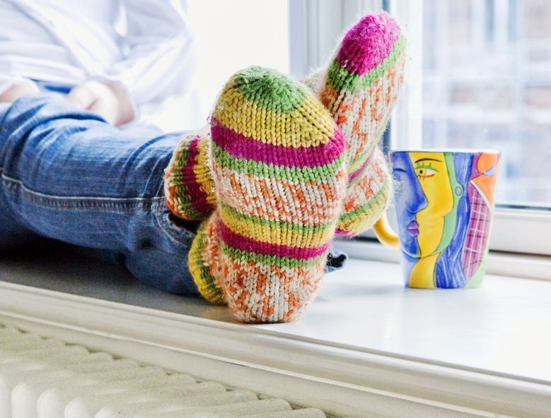 Mann som sitter i en vinduskarm med fargerike ullsokker og en kopp med varmt drikke. Foto.