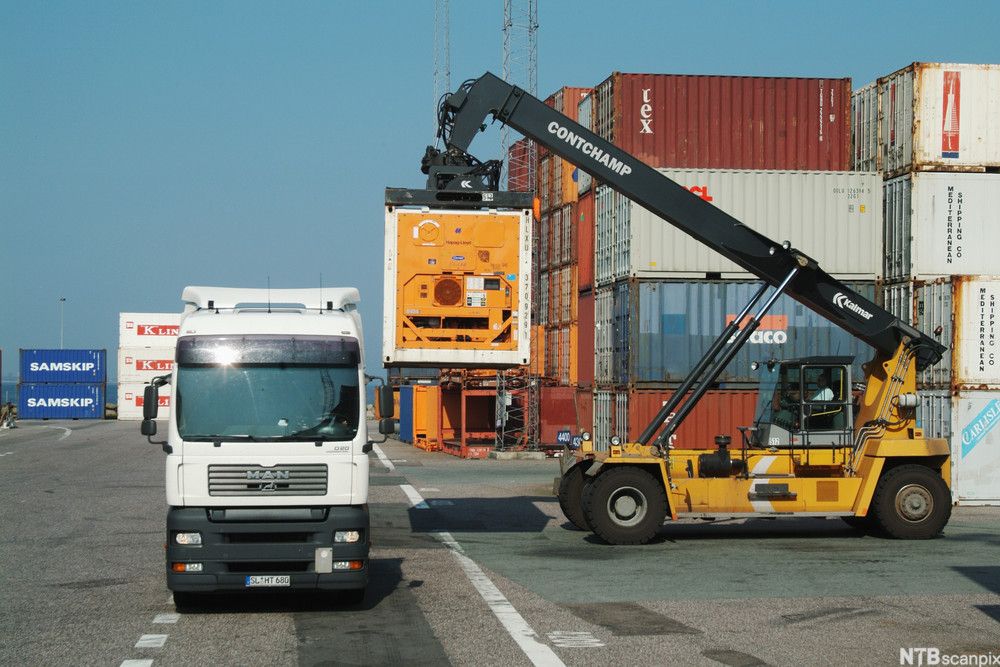Bildet viser en truck som laster en termokonteiner på en lastebil på et havneområde. Foto.