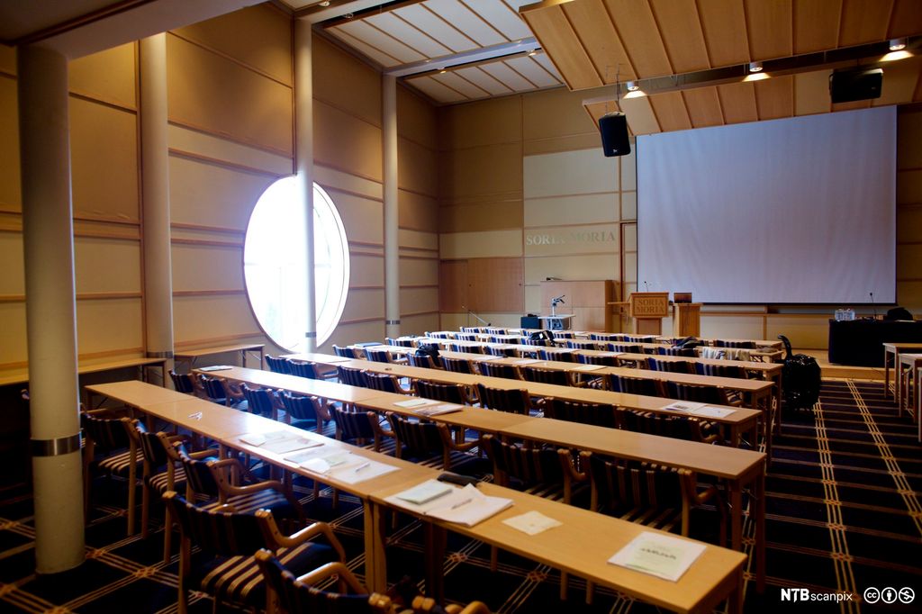 Tomt konferanserom  med seks ganske breie, todelte benkerader. Stort lerret bak talerstolen, og både mikrofoner og annet lydutstyr. Foto.