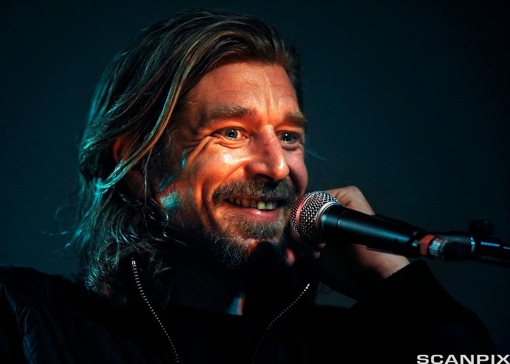 Portrett av ein mann med skjegg og halvlangt hår som smiler framfor ein mikrofon. Foto.