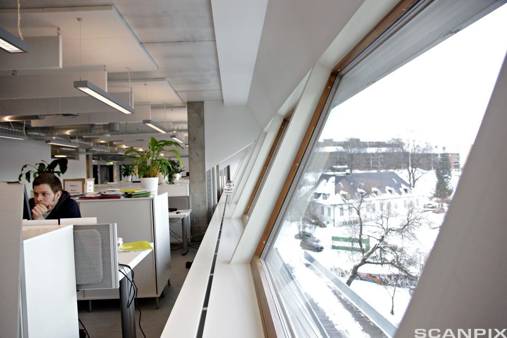 Mann sitter ved pulten sin i et kontorlandskap. Vegg med vinduer til høyre. Foto.