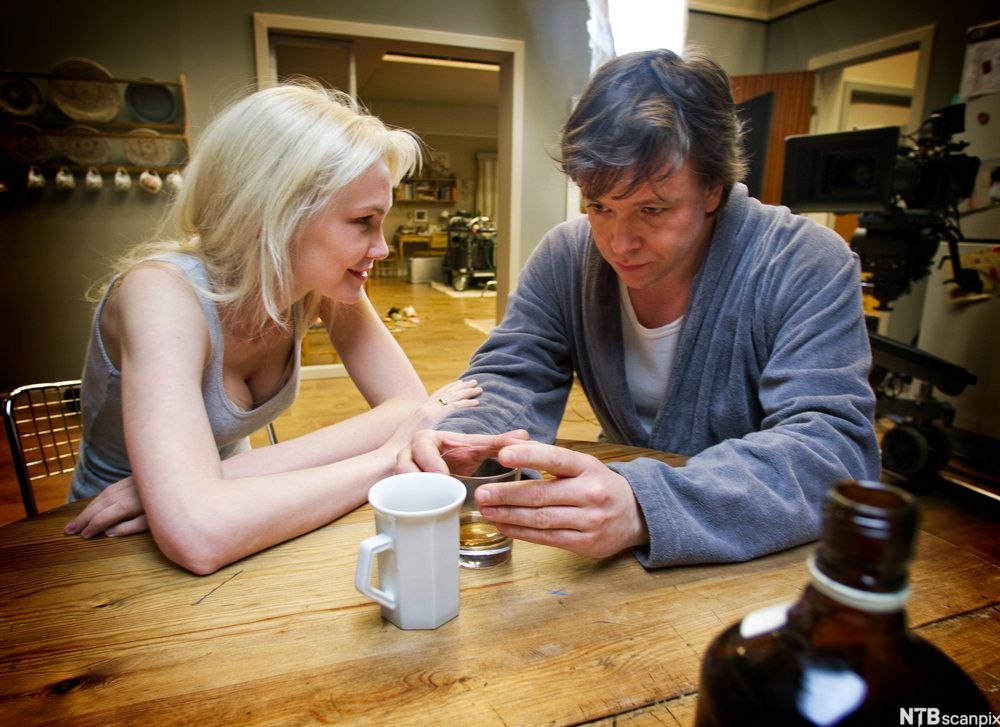 Filmscene med en ung kvinne og en ung mann ved et bord. Mannen holder et whisky-glass. Foto.