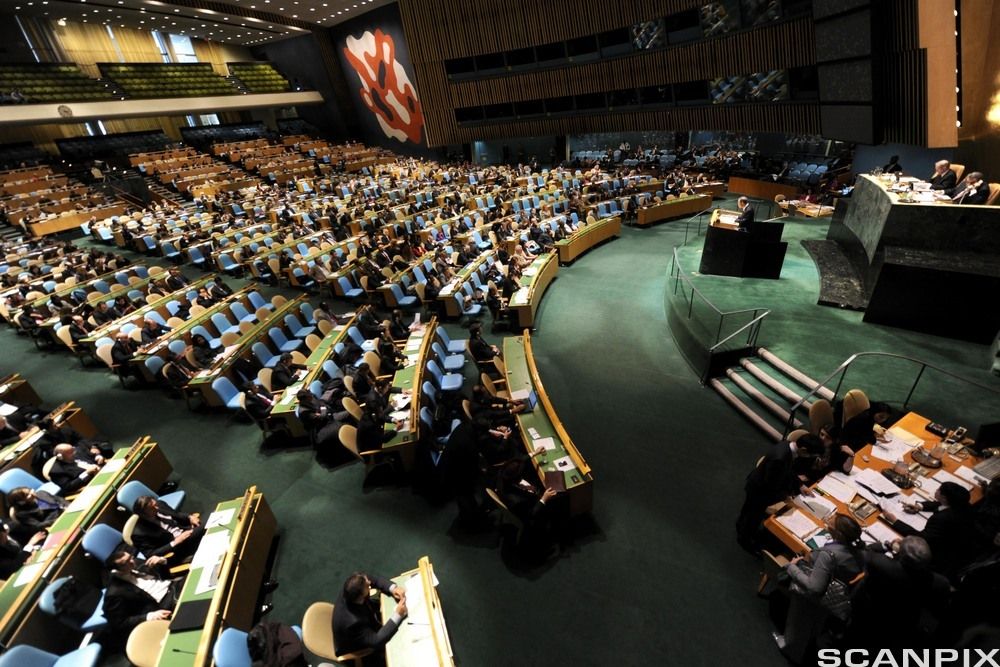 Stor sal med mange rader med sitteplasser. Til høyre et podium. Foto.