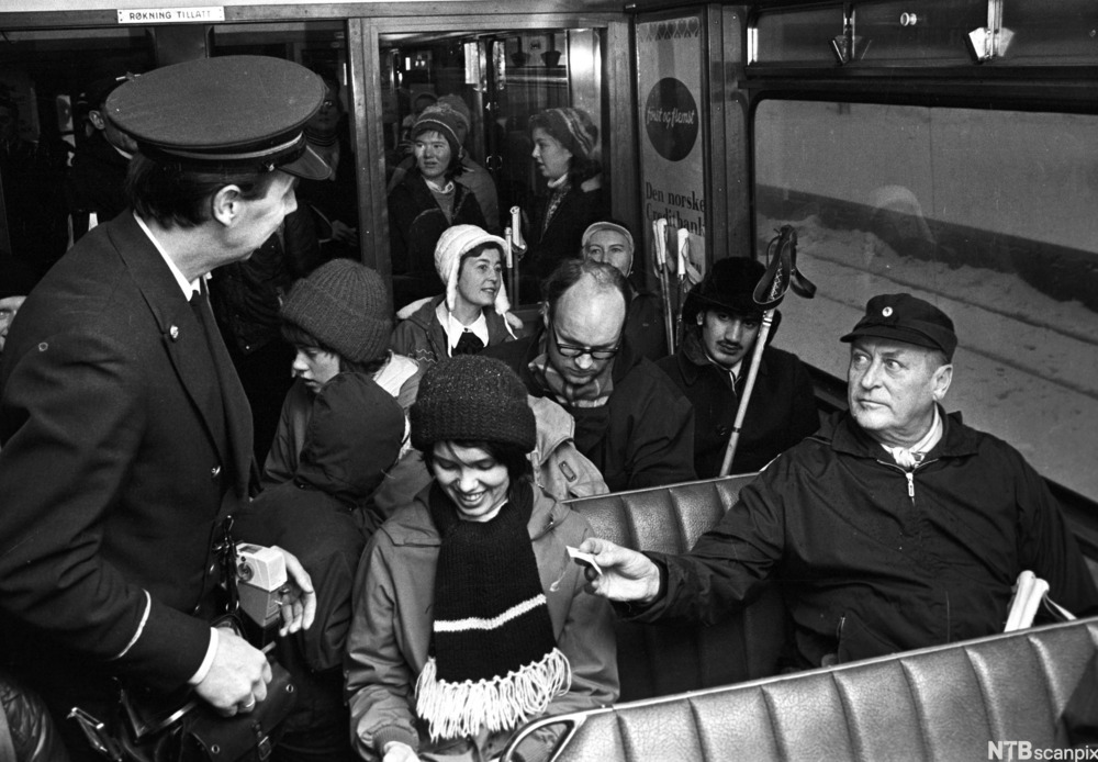 Kong Olav, en eldre mann i anorakk og skyggelue, sitter på en vindusplass i en trikk og rekker konduktøren billetten sin. En ung kvinne sitter ved siden av kongen og ler beskjedent mens hun ser ned. Flere passasjerer med skistaver i bakgrunnen. Foto.