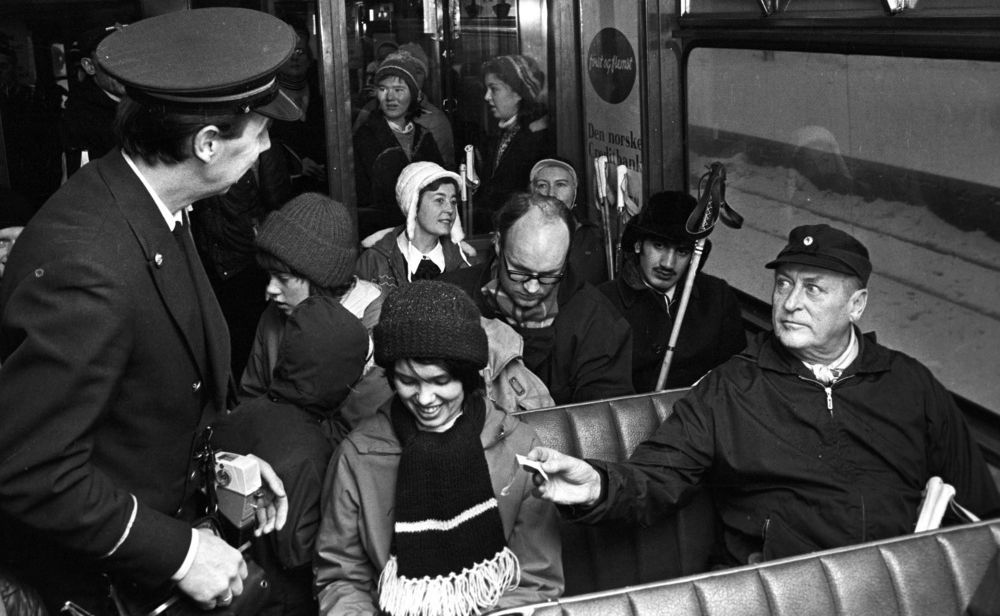 Kong Olav, en eldre mann i anorakk og skyggelue, sitter på en vindusplass i en trikk og rekker konduktøren billetten sin. En ung kvinne sitter ved siden av kongen og ler beskjedent mens hun ser ned. Flere passasjerer med skistaver i bakgrunnen. Foto.