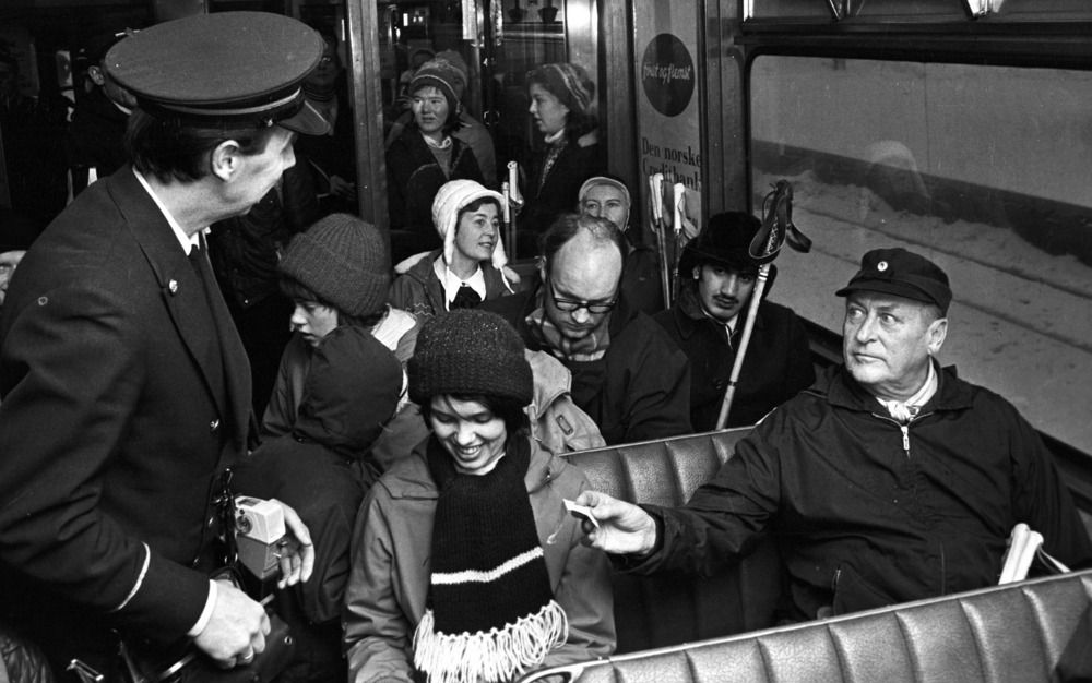 Kong Olav, ein eldre mann i anorakk og skyggelue, sit på ein vindaugsplass i ein trikk og rekker konduktøren billetten sin. Ei ung kvinne sit ved sida av kongen og ler beskjedent mens ho ser ned. Fleire passasjerar med skistavar i bakgrunnen. Foto.
