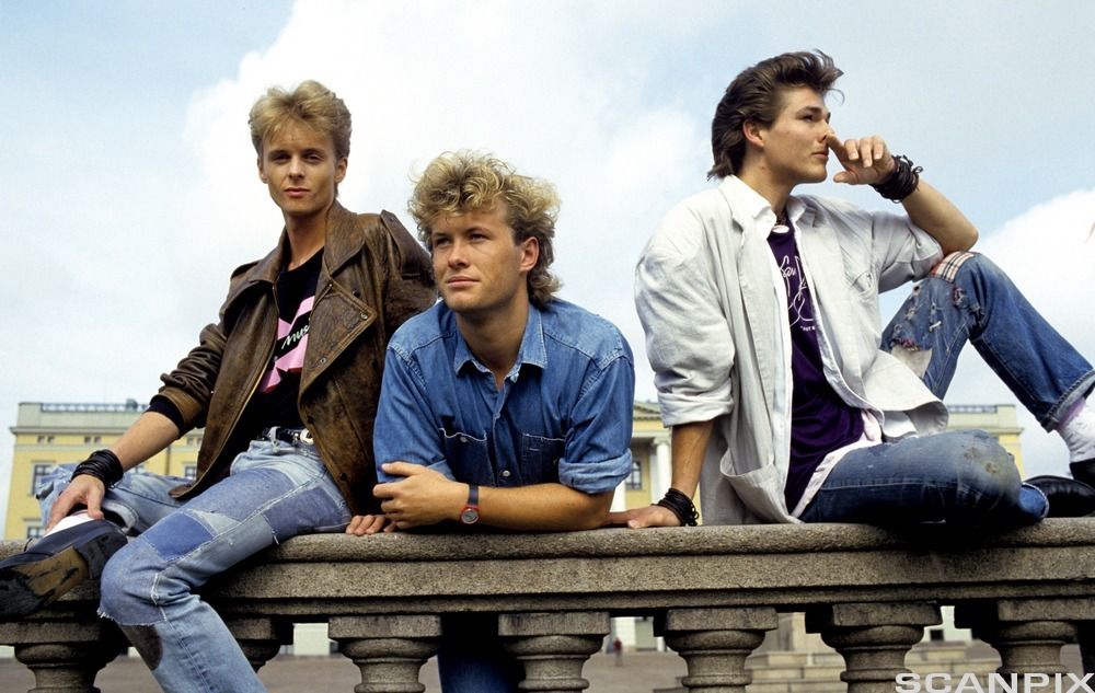 Paal Waaktaar, Magne Furuholmen og Morten Harket sit på og står ved eit gjerde mens dei ser i forskjellige retningar. Foto. 