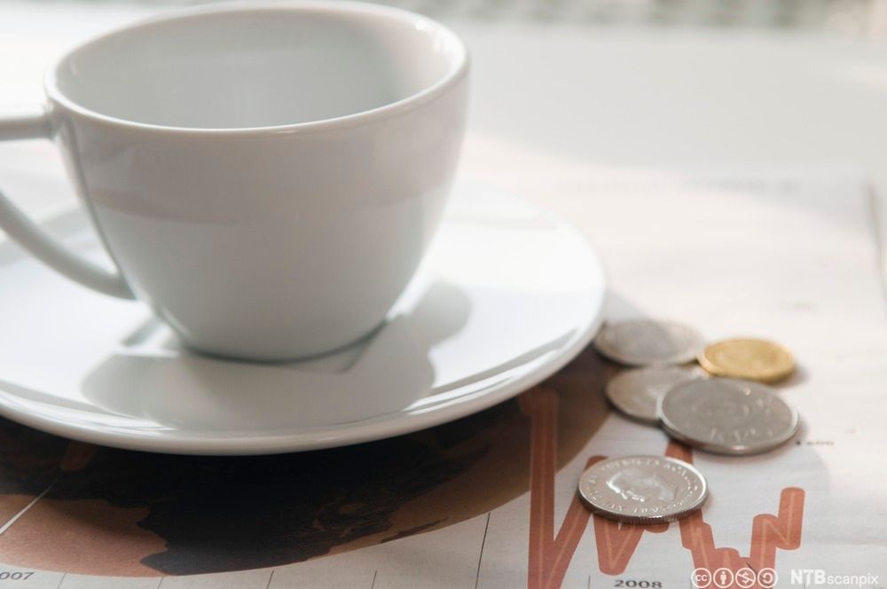 En hvit kaffekopp med skål. Ved siden av ligger noen mynter. Foto.