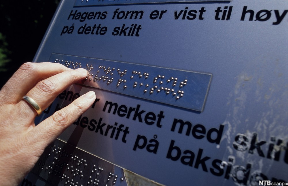 Hånd leser punktskrift på ei informasjonstavle. Foto.