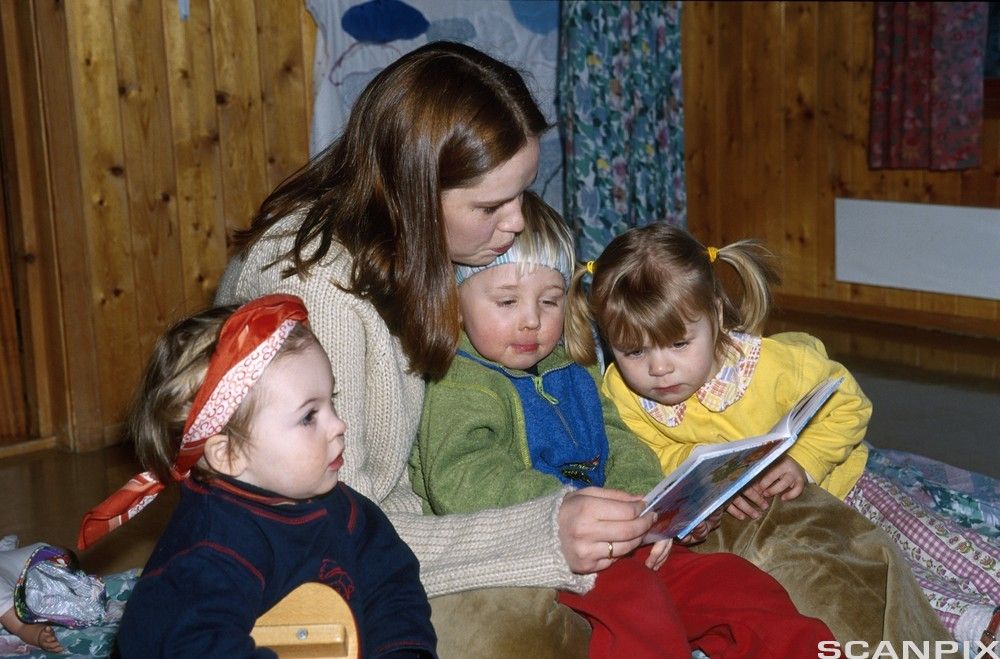 En voksen dame sitter på gulvet og leser ei bok for tre barn.