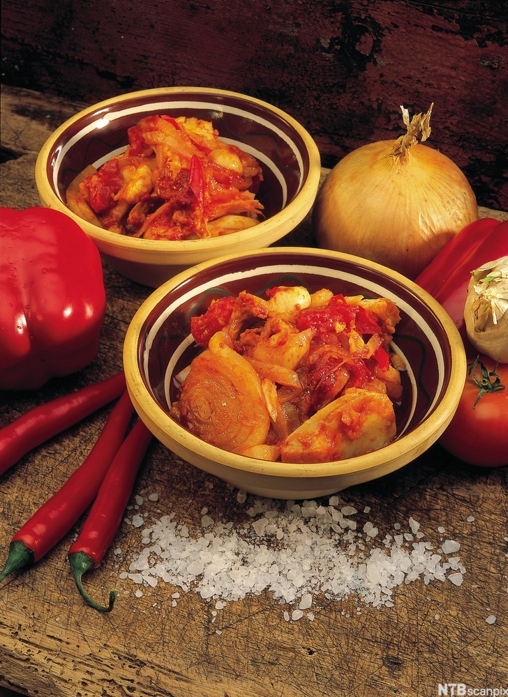 En porsjon bacalao i en skål. Noen hele grønnsaker, blant annet paprika, løk og chili, ligger ved siden av. Foto.
