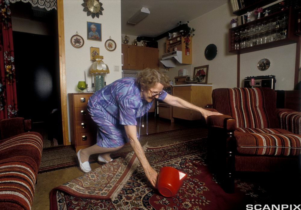 Eldre dame snubler i et gulvteppe. Foto.