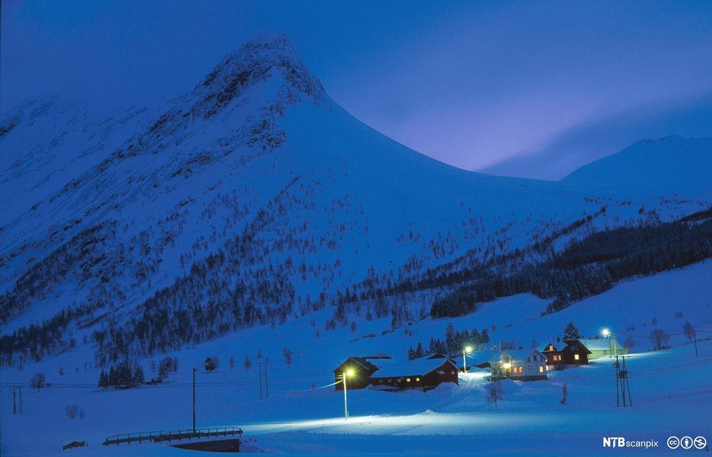 Oversiktsbilde over snødekt vinterlandskap. Fjelltopper i bakgrunnen og en større gård med opplyste vinduer lenger framme. Foto.