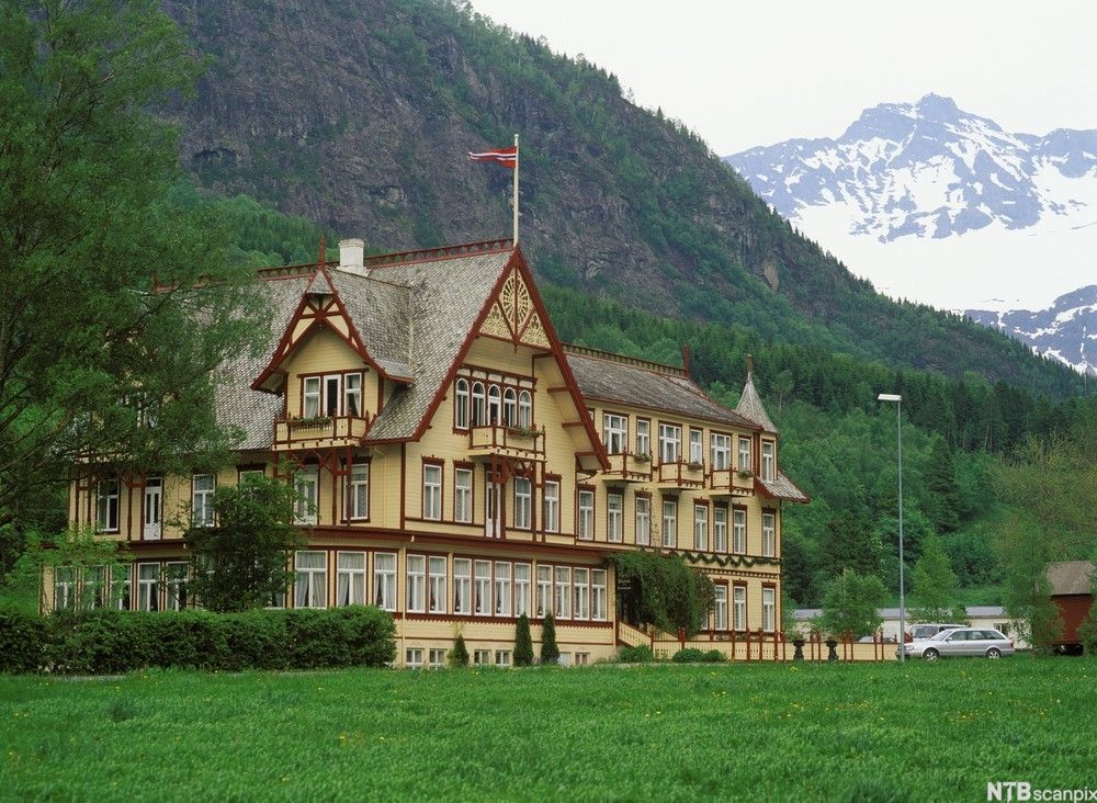 Ærverdig trehus i sveitserstil i et grønt og frodig landskap med mektige, snødekte fjell i bakgrunnen. Foto.