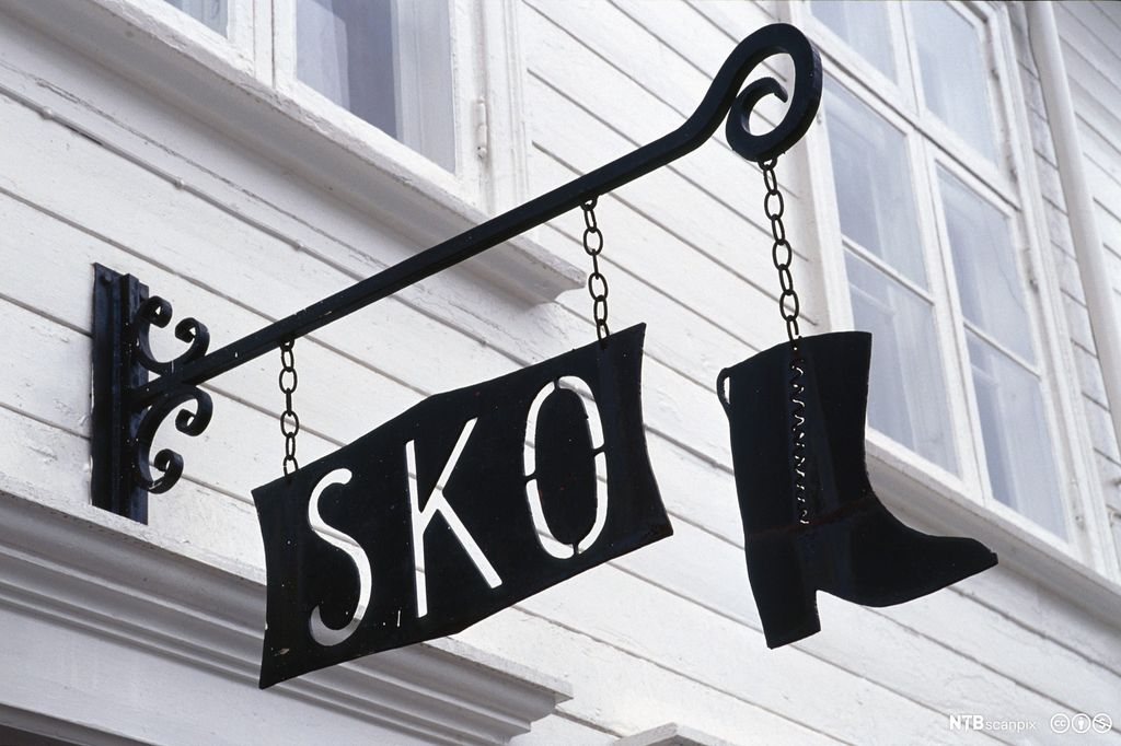 En svart støvlett og et svart skilt hvor det står ordet "sko", henger over døra til en skobutikk. Foto.