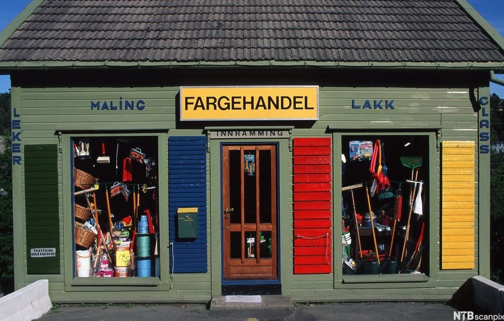 Grønnmalt trehus som huser en liten, gammeldags fargehandel. Noen felter på fasaden er malt i mørkegrønt, blått, rødt og gult, og utstillingsvinduene er overfylt med varer. Foto.