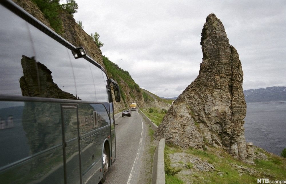 En turistbuss på en smal vei langsmed en innsjø. Foto.