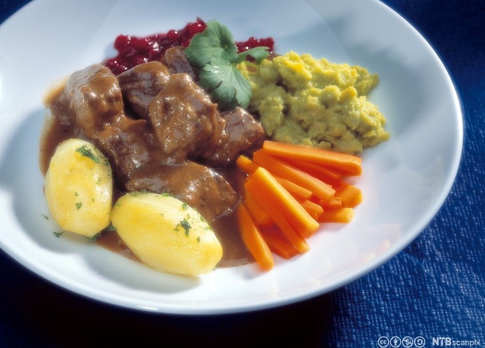 Småsteik av okse, ertestuing, kokte grønnsaker, kokte poteter og tyttebærsyltetøy servert på en hvit middagstallerken. Foto.