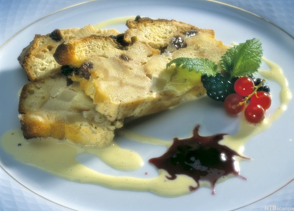 Brødpudding basert på to skiver brød delikat anrettet på en blå tallerken. Striper med vaniljesaus går i sirkler rundt skivene. En håndfull bær, et lite grønt blad og en klatt mørk kompott er tilbehør på tallerkenen. Foto.