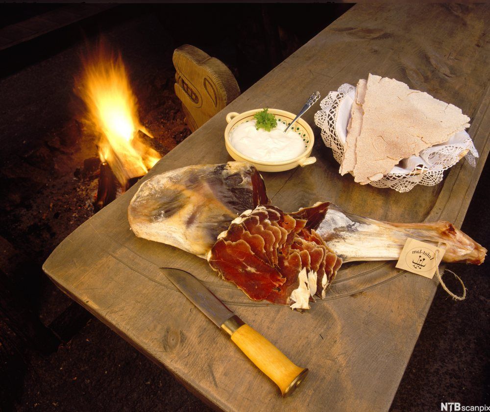 Fenalår tilberedt med rømme og flatbrød på ei fjøl foran en peis med flammer. Foto.