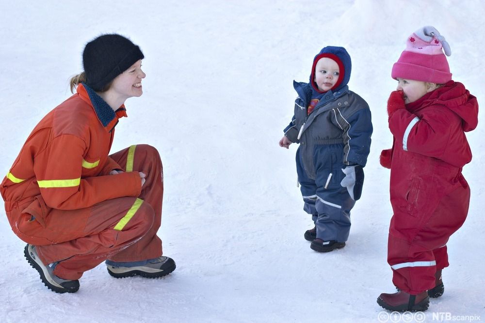 Barnehagetilsett pratar med to barn. Alle tre har på seg vinterdressar. Den vaksne sit på huk slik at ho har ansiktet i same høgde som barna. Foto.
