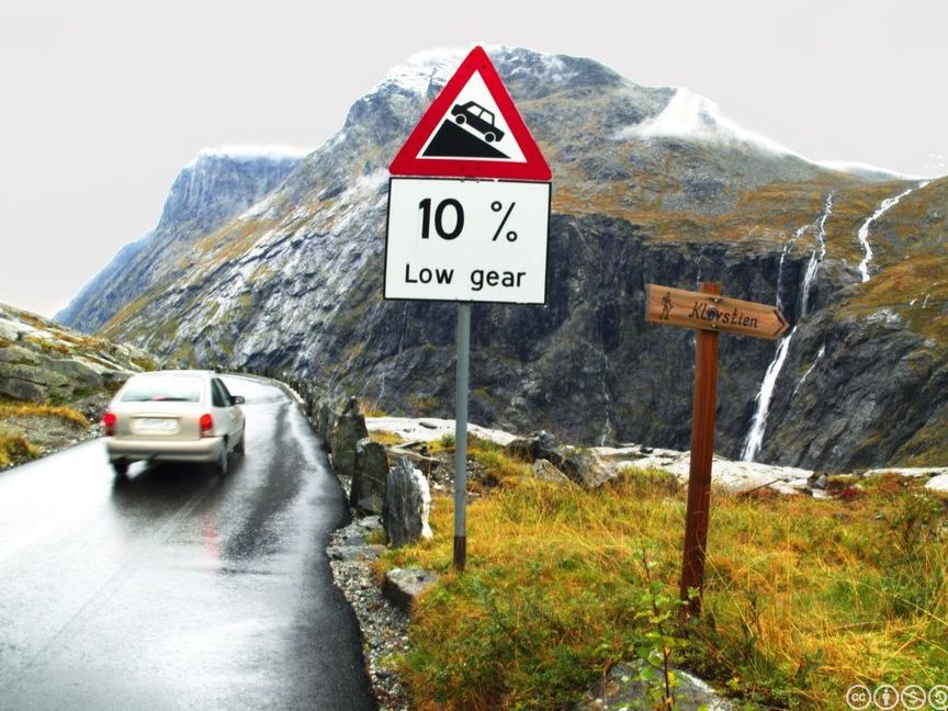Ein bil er på veg til å køyre ned ein bratt bakke i eit fjellandskap. Foto.