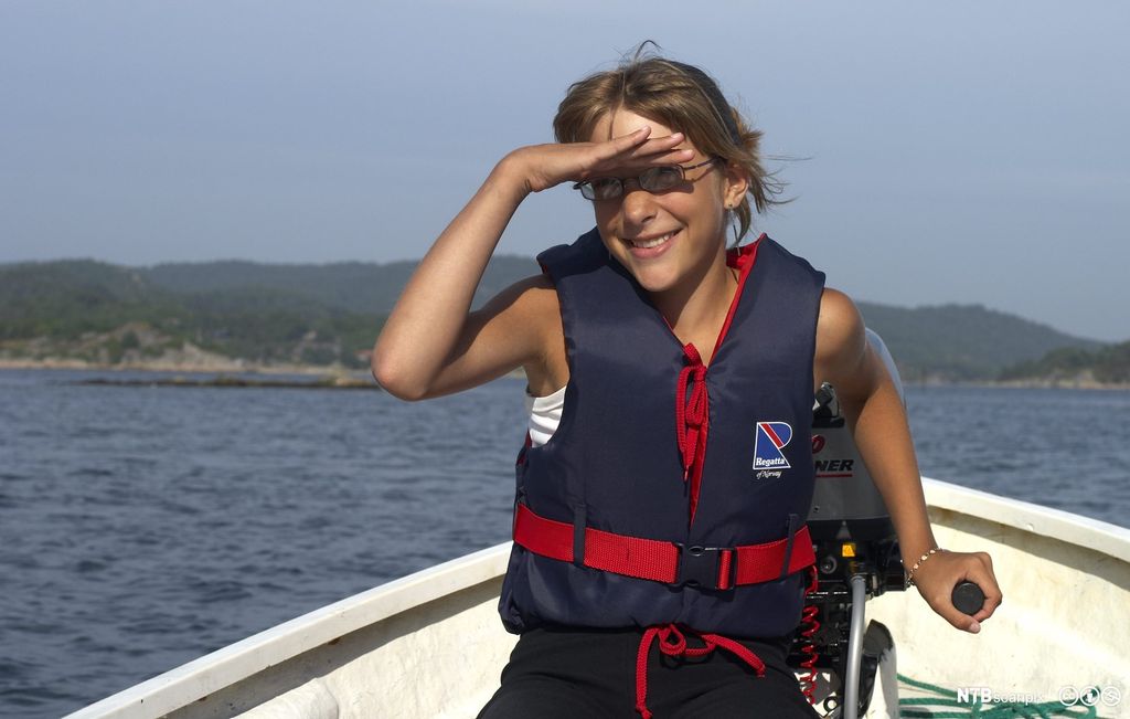 Jente med flytevest styrer en liten båt med påhengsmotor. Foto.