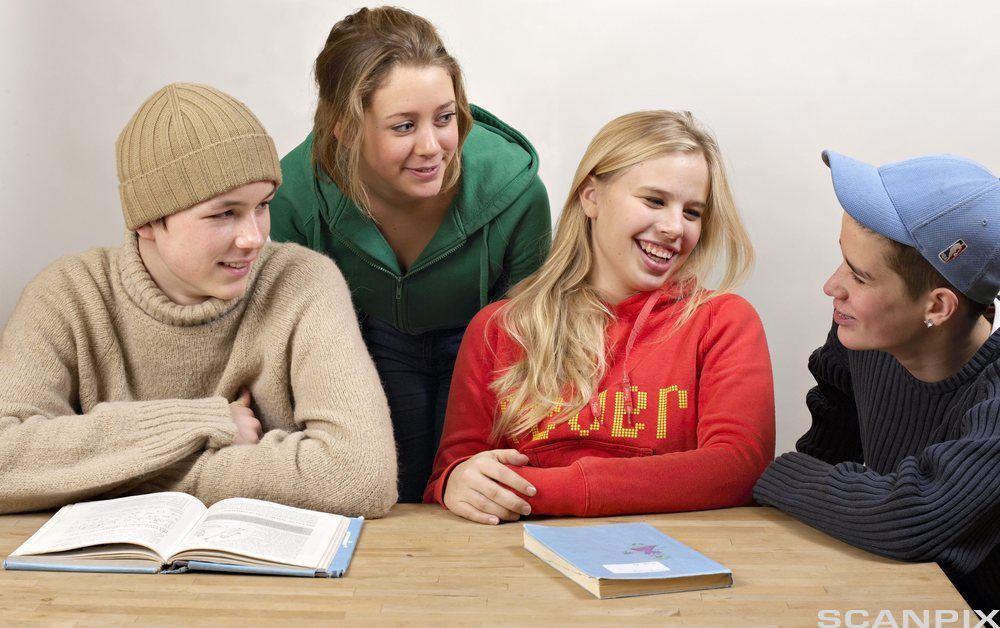 Fire ungdommer i godt humør sitter ved et bord med ei lukket og ei oppslått bok foran seg. De ser på hverandre og smiler. Foto.