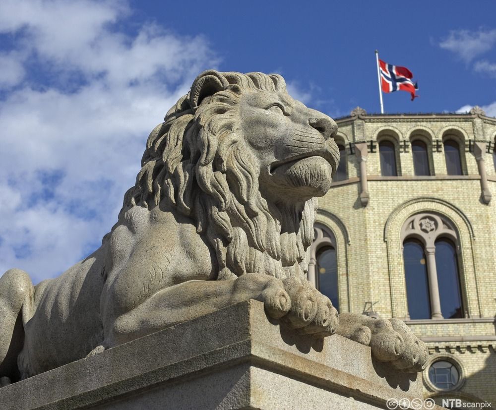 En statue i stein av en liggende løve. I bakgrunnen vaier det norske flagget på toppen av en murbygning. Foto.