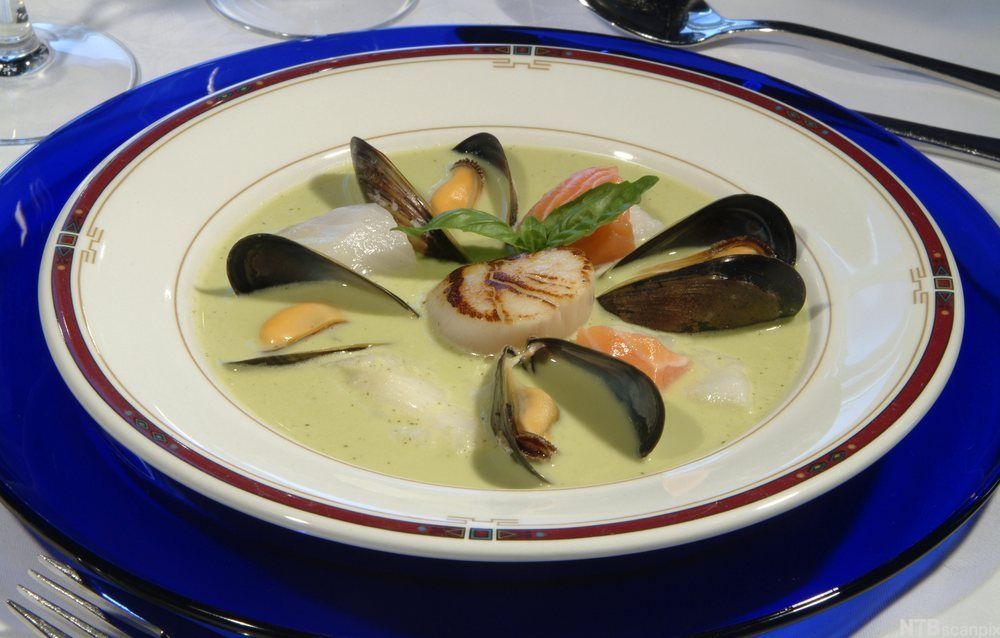 Skål med lys suppe med blåskjell, andre skjell og hvit og rød fisk. Foto. 