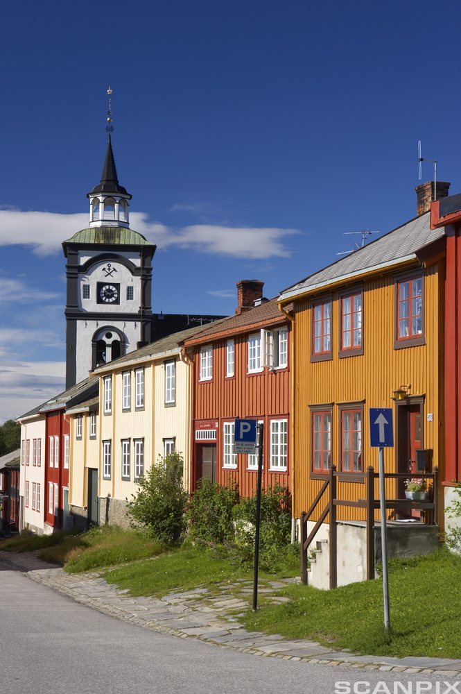 Svart og hvitt kirketårn mot blå himmel med lavere hus i forgrunnen. Foto.