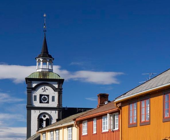 Svart og hvitt kirketårn mot blå himmel med lavere hus i forgrunnen. Foto.