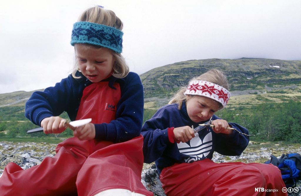 To jenter med røde regnbukser bruker kniv og spikker på hver sin pinne. Foto.