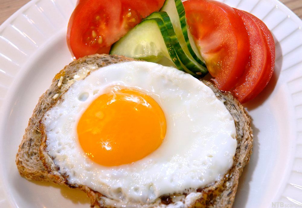 Speilegg med hel plomme på ei grov brødskive med tomat og agurk ved siden av. Foto.